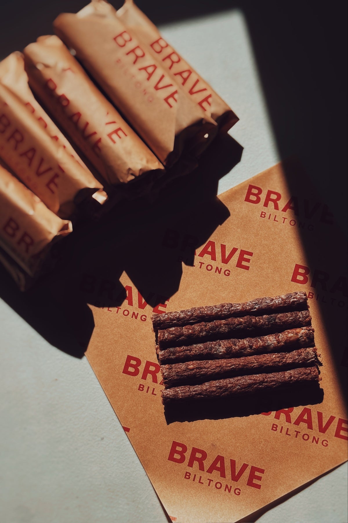 Brave Biltong meat sticks arranged on branded brown paper packaging with several wrapped sticks in the background, photographed in dramatic side lighting on a light surface.