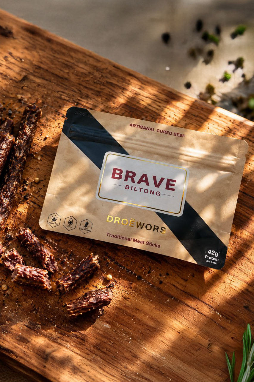 A package of Brave Droëwors traditional meat sticks lies on a rustic wooden board with some sliced pieces beside it. The package is metallic gold with a diagonal black stripe and a central white label reading 'BRAVE BILTONG'.