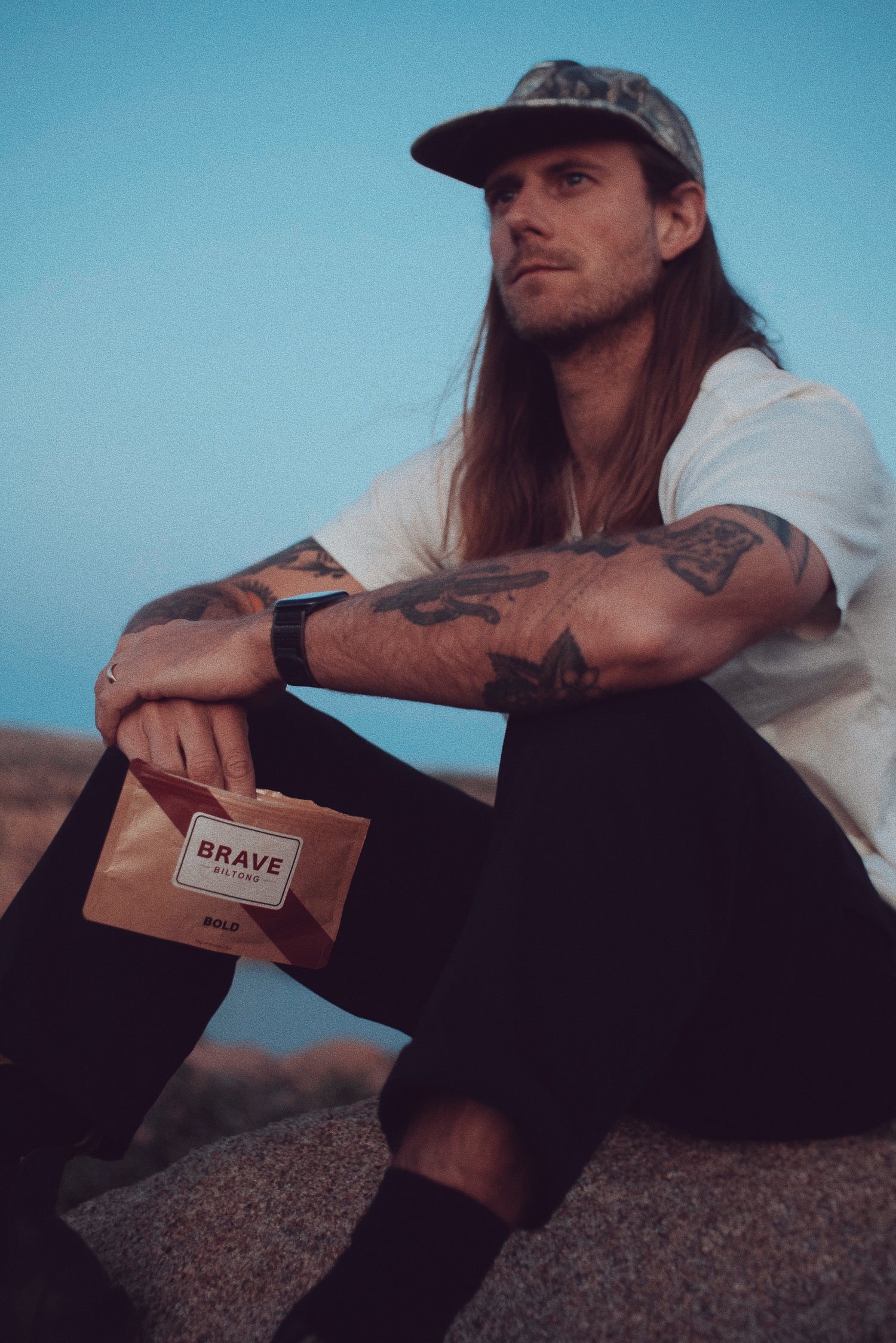 Man with tattoos holding a 'Brave Biltong' branded package against a blue sky.